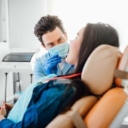 A dentist wearing a mask and gloves examines a patient sitting in a dental chair in a brightly lit clinic.