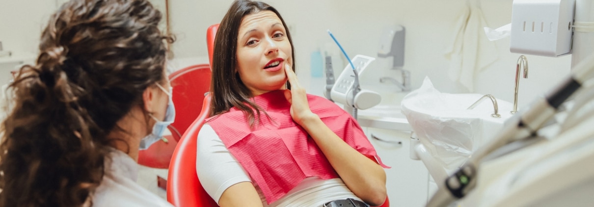 A woman sits in a dental chair holding her jaw and appears to be in discomfort while a dentist speaks to her.