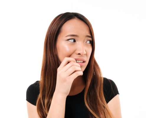 A woman with long brown hair and a black shirt looks to the side with a worried expression, biting her nails against a white background.