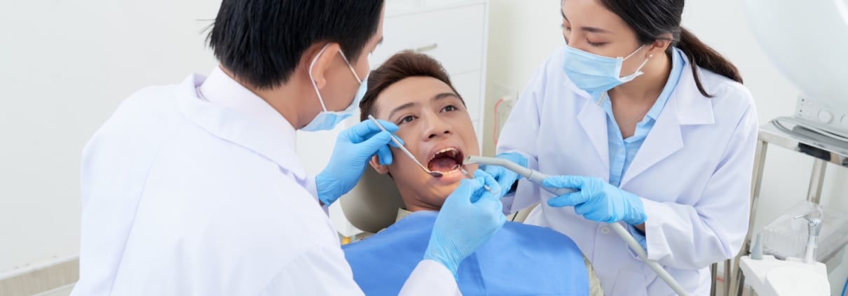A dentist examines a patients mouth with dental tools while a dental assistant assists, both wearing masks and gloves in a dental clinic.