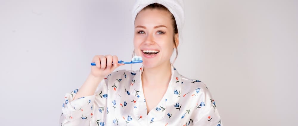 Woman in pajamas and a towel wrapped around her head is brushing her teeth and smiling, standing against a plain light background.