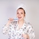 Woman in pajamas and a towel wrapped around her head is brushing her teeth and smiling, standing against a plain light background.