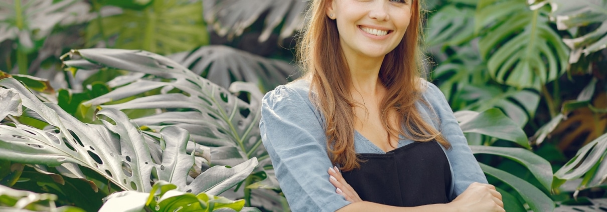 A woman wearing a black apron stands with arms crossed, smiling in front of large green tropical plants.
