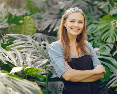 A woman wearing a black apron stands with arms crossed, smiling in front of large green tropical plants.