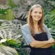 A woman wearing a black apron stands with arms crossed, smiling in front of large green tropical plants.