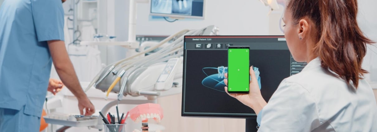 A dental professional holds a smartphone with a green screen in front of a computer displaying dental X-rays, while another person works in the background.