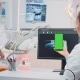 A dental professional holds a smartphone with a green screen in front of a computer displaying dental X-rays, while another person works in the background.