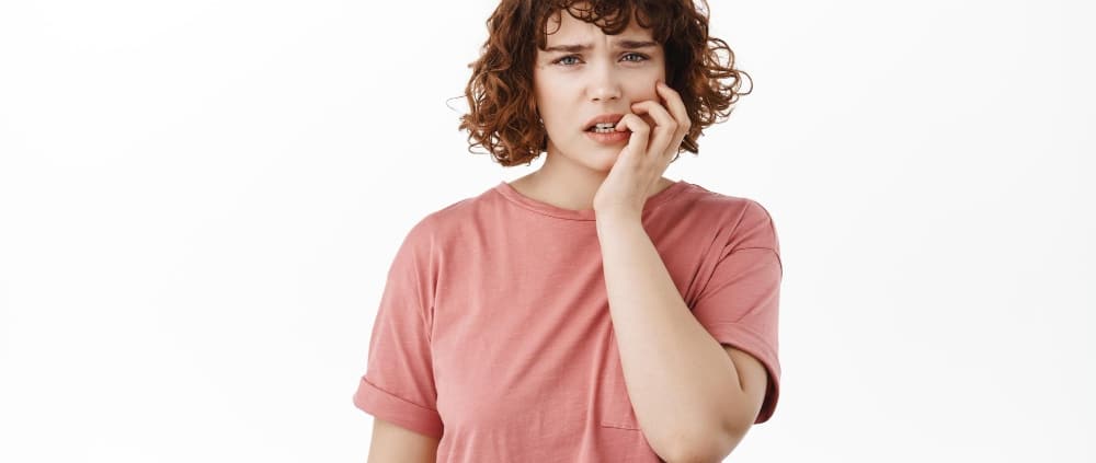 A young woman with curly hair wearing a pink t-shirt stands against a white background, looking concerned and touching her face with one hand.