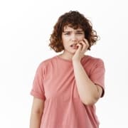 A young woman with curly hair wearing a pink t-shirt stands against a white background, looking concerned and touching her face with one hand.