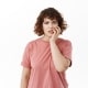 A young woman with curly hair wearing a pink t-shirt stands against a white background, looking concerned and touching her face with one hand.