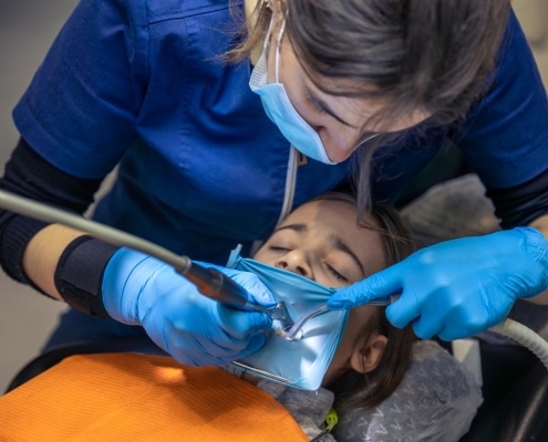 A dentist wearing blue gloves and a mask performs a dental procedure on a young patient lying in a dental chair.
