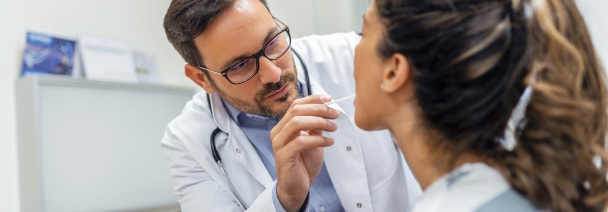 A doctor wearing glasses and a white coat examines a patients throat with a tongue depressor in a medical office.