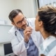 A doctor wearing glasses and a white coat examines a patients throat with a tongue depressor in a medical office.