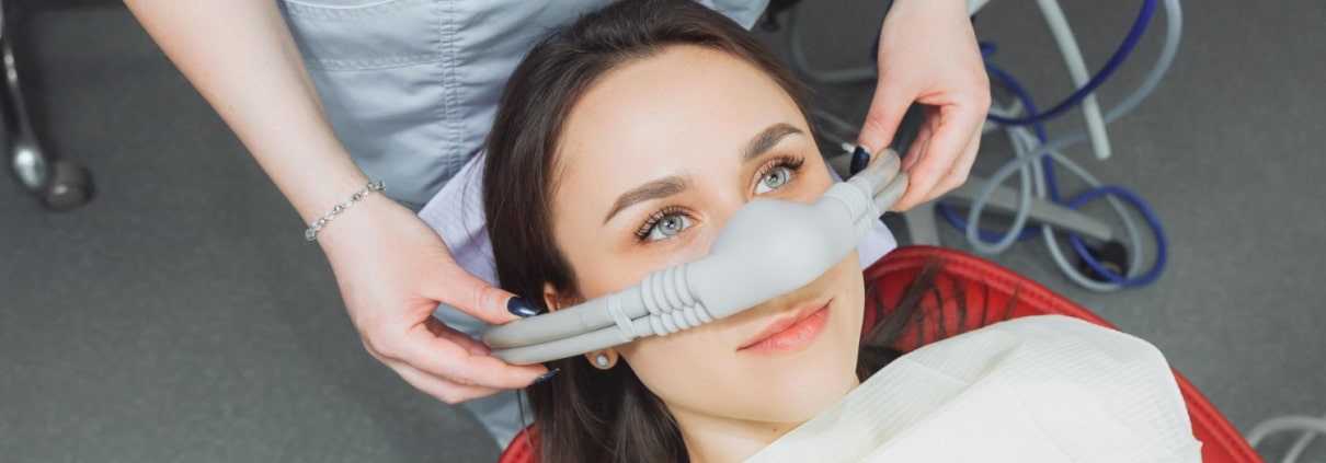 A dental patient reclines in a chair while a dental professional places a nitrous oxide sedation mask over her nose.