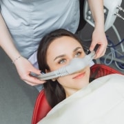 A dental patient reclines in a chair while a dental professional places a nitrous oxide sedation mask over her nose.
