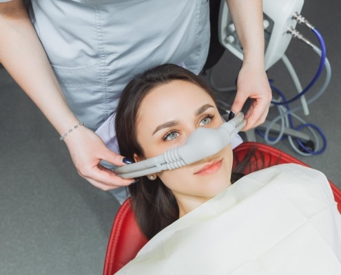 A dental patient reclines in a chair while a dental professional places a nitrous oxide sedation mask over her nose.